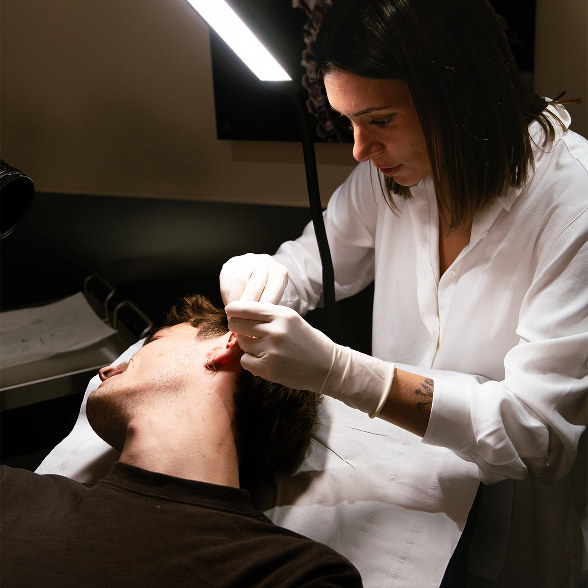 Perceuse en blouse blanche réalisant un piercing d'oreille sur un client allongé en cabinet